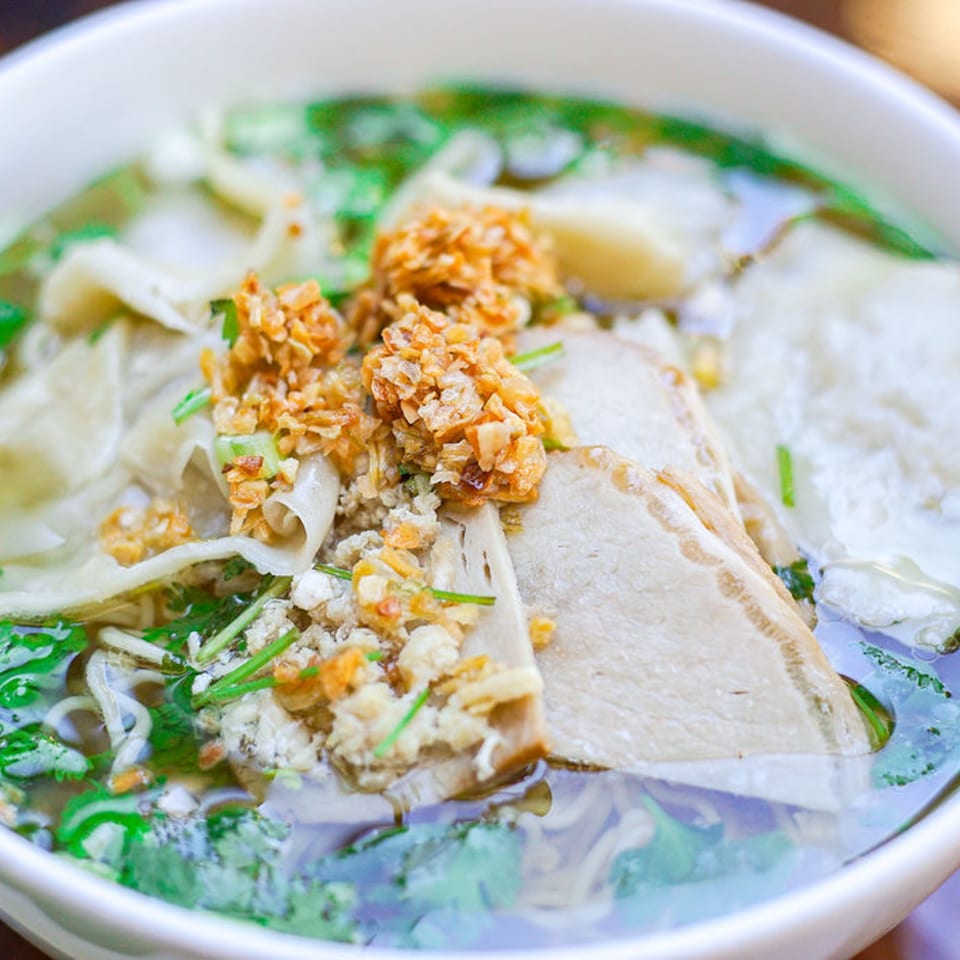 Close-up of a Vietnamese noodle soup with meat, herbs, and crispy fried garlic in a white bowl.