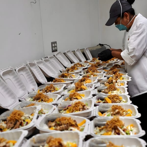 A chef in a mask and gloves prepares numerous takeout containers of Vietnamese food with crispy toppings.